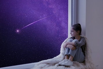 Cute little girl sitting on windowsill and looking at shooting star in beautiful night sky