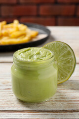 Jar of avocado dip, lime and plate with french fries on white wooden table