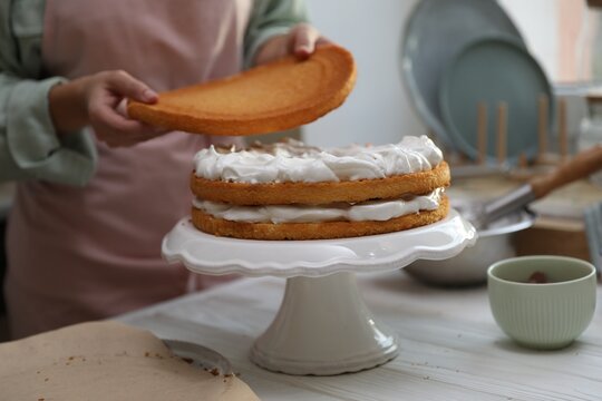 Woman stacking layers of sponge cake at white wooden table in kitchen, closeup
