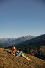 Woman full-length sitting resting on a hill and looking at the mountains in a yellow raincoat and jeans happy camping trip in the winter, freedom lifestyle 