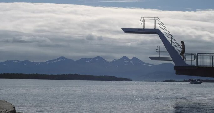 Wide scenic shot of long haired, blonde, caucasian male climbing and jumping from diving tower doing a backflip gainer into water in profile in Norway