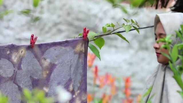 an Asian Muslim woman, wearing a hijab is drying cloth among the leaves, the background is blurred.