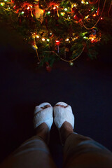 female legs in white fluffy cozy slippers near a festive Christmas tree in the evening light of lights on a dark background, top view - 553626518