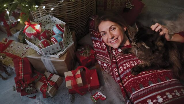 A cute girl is lying under the Christmas tree covered with gifts and enjoying the company of her cat. She is dressed in beautiful Christmas clothes. Slow motion. Concept of Christmas celebration