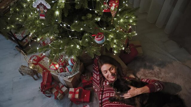 A cute girl is lying under the Christmas tree covered with gifts and enjoying the company of her cat. She is dressed in beautiful Christmas clothes. The concept of the New Year's celebrating.