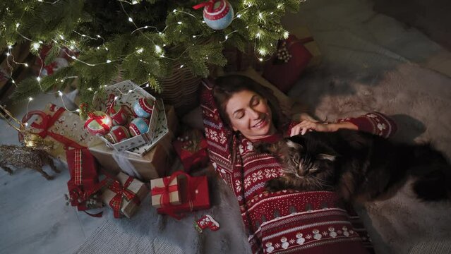 A beautiful girl dressed in New Year's clothes lies under the Christmas tree in the company of her favorite cat. Camera move away. The concept of the New Year's celebrating.
