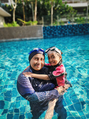 Fototapeta na wymiar Smiling mother and her adorable little daughter hugging each other while standing together in a swimming pool at a tropical resort