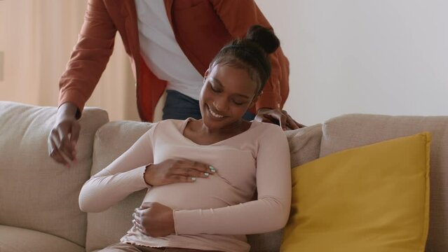Young happy pregnant black woman resting on sofa, her loving husband coming and caressing big belly, empty space