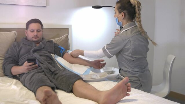 Patient lying in a hospital bed and scrolling on his smartphone while young beautiful nurse is setting up the needle and IV stand for intravenous therapy