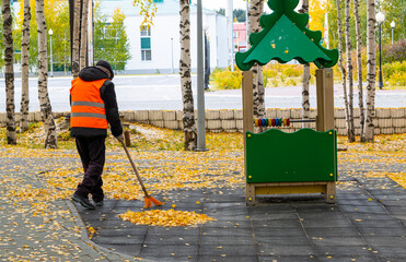A man sweeps yellow leaves in an autumn park on a playground