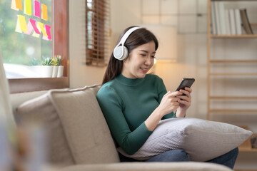 Beautiful Asian woman sitting on sofa in living room using mobile phone playing games and various entertainment programs at home holiday and having fun on vacation.