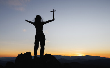 Silhouette of christian young woman praying with a  cross at sunrise, Christian Religion concept background.