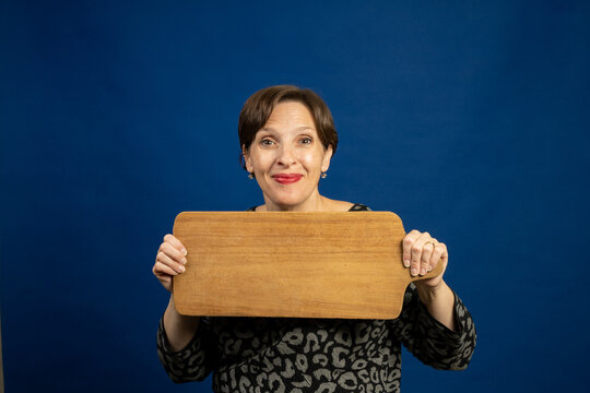 Happy Older Woman Holding A Cutting Board As A Sign. High-quality Photo With Room For Copy On Wooden Board