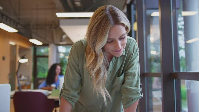 Camera pulls back to show mature businesswoman standing at desk in office approving layouts or proofs and drinking coffee - shot in slow motion