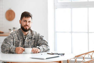Young soldier in wheelchair with cup of coffee at table in kitchen