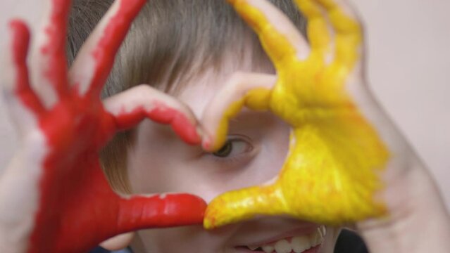 portrait of happy caucasian boy looking through painted hands showing heart shaped gesture.selective focus.boy with painted palms looking at camera