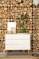 Chest of drawers with houseplant, blank frames and basket near wooden wall