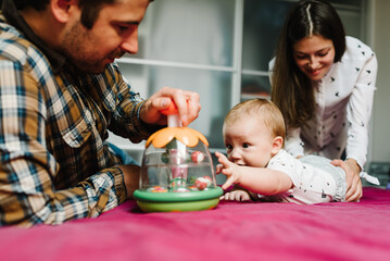 Happy family, parenthood and people concept - mother, father with baby lying in bed at home. Portrait of young smiling family with son playing with toy.