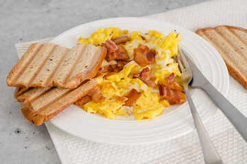 Scrambled eggs fried with cheese, and served with fried bacon and toasted bread close-up on a white plate