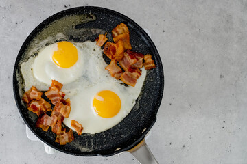 Fried eggs and crispy fried bacon slices close-up on a frying pan, view from above, copy space.