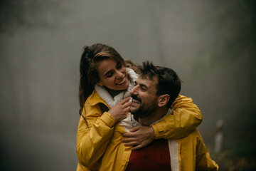 Loving couple with raincoats having a great time, hugging and having a piggyback ride on a cold day in the forest.