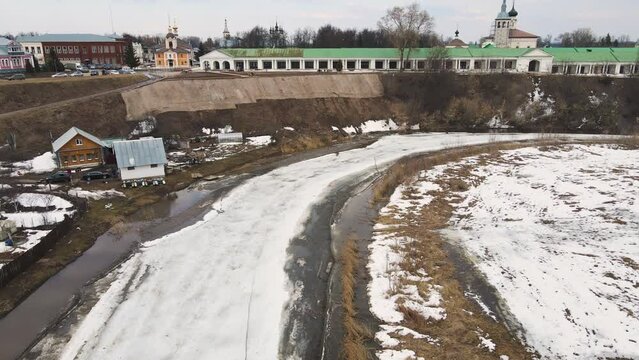 A curved small frozen river, the ice is melting, there is a town on the bank. Spring, snow melts, dry grass, flood and river flood. Day, cloudy weather, soft warm light. UHD 4K