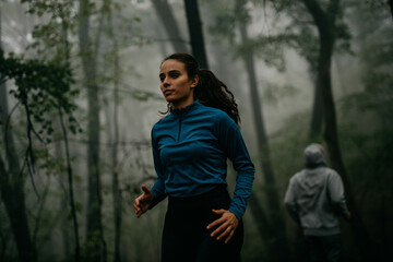 Focused and determined woman and man having a sports recreation in the foggy forest. Focus on a woman in a blue sports outfit, a man in the background.
