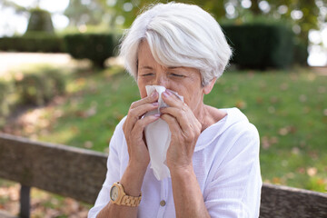 senior woman with tissue having flu or allergy at park - obrazy, fototapety, plakaty