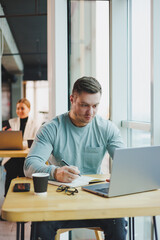 Fototapeta premium A young male manager sits in an office with large windows and works at a laptop. A handsome businessman is working on his homework in a cafe.