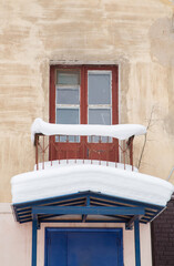 A small balcony covered with snow over a canopy with a large white snowdrift. Glazed wooden old door, a balcony above the entrance to the entrance. Winter cloudy day, soft light.