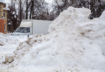 A huge, dirty snowdrift against the backdrop of a city street with cars. On the road lies white snow in high heaps. Urban winter landscape. Cloudy winter day, soft light.