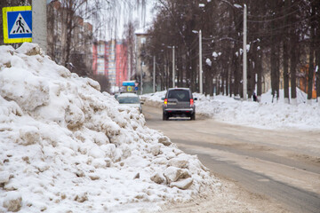 A large, high snowdrift against the backdrop of a city street with cars. On the road lies white snow in high heaps. Urban winter landscape. Cloudy winter day, soft light.