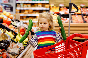 Cute todler girl pushing shopping cart in supermarket. Little child buying fruits. Kid grocery shopping. Adorable baby kid with trolley choosing fresh vegetables in local store.