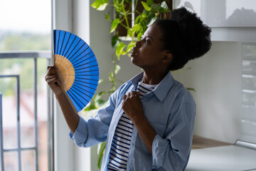 Young casual African American woman fanning herself with hand fan experiencing health problems due...