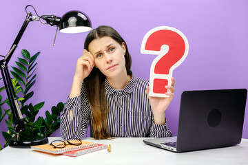 Pensive sad young employee woman 20s old years sitting work at white office desk pc laptop holding red question mark sign, posing isolated over purple color background wall in studio. Business concept