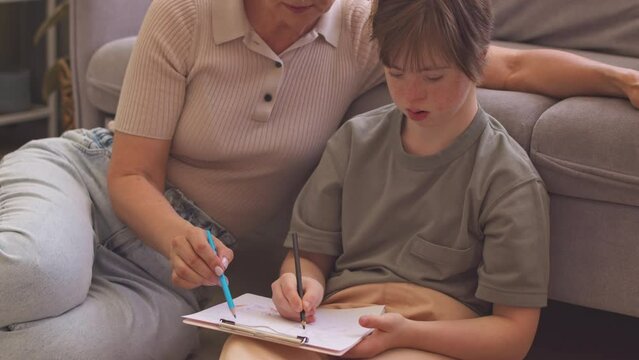 Concentrated girl with down syndrome and her caring mother sitting together in living room drawing