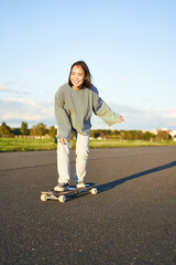Cute asian girl riding skateboard, skating on road and smiling. Skater on cruiser longboard...