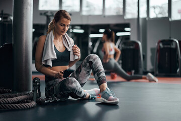 Pretty woman working out in a gym, making pause and using smartphone. Adult pretty sporty lady with...