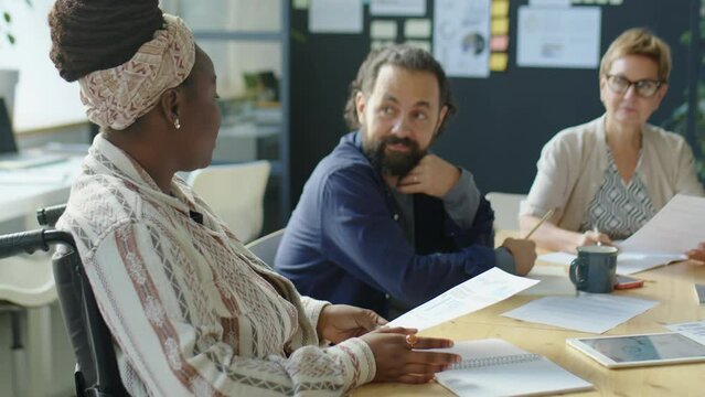 Tilt up shot of African American woman in wheelchair discussing business paper with multiethnic coworkers during team meeting in office