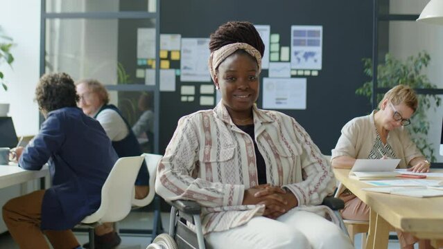 Medium long shot of young African American businesswoman with disability sitting in wheelchair and posing for camera with smile during workday in open space office