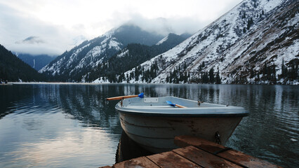 A mountain lake in the forest with mirrored water. Wooden pier with white boat. The water reflects the landscape of a cloudy sky, snowy mountains and peaks, coniferous firs. Kolsai Lake, Kazakhstan