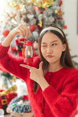 Young beautiful friendly asian female lady with red long sleeve sweater shirt and cute headband cheerfully holding a small lantern light beside a nicely decorated Christmas tree in a room