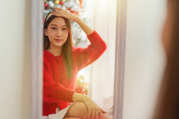 Portrait photo of a young beautiful friendly asian female lady in red long sleeve sweater and white skirt with nice headband posing different looks in front mirror with a nice decorated Christmas tree