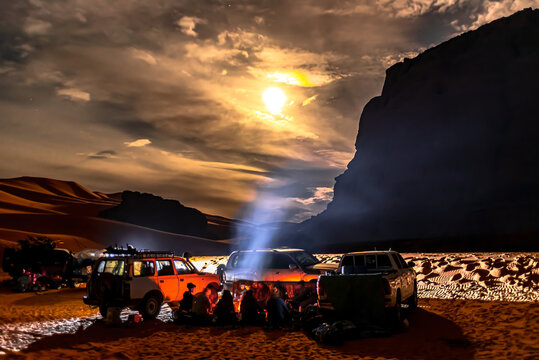 Campfire group and cars in Sahara Desert by night. 4X4 vehicules parked, Unrecognizable tourists and local berber people sitting around a camp-fire in Tadrart Rouge. Cloudy sky and full moon moonlight