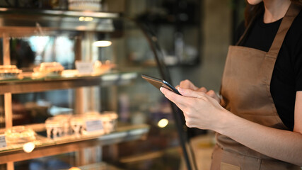 Cropped shot of woman coffee shop entrepreneur wearing an apron leaning against counter and using a digital tablet