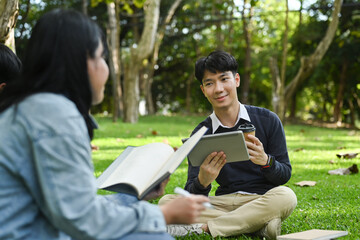 Smiling young asian male checking university schedule on digital tablet and talking with his friend while sitting on green grass in campus