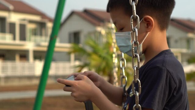 Asian kid wear face mask play mobile phone at swing in playground at suburban town