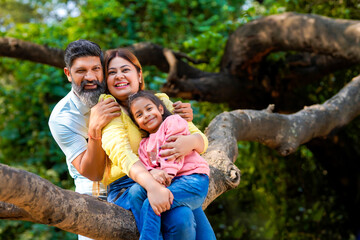 Naklejka na ściany i meble Happy indian couple sitting with his little girl on tree branch at garden.