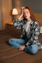 Vertical portrait of sick ginger woman with closed eyes touch head, suffering from awful headache, sitting on bed at home. Young tired female feeling unhealthy, strong sudden pain, morning hangover.