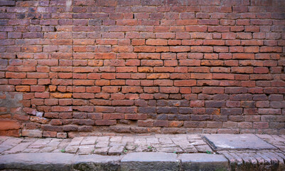 Frontal view of an old red-brown brick wall in a back street of Kathamandu. In the foreground a gray cobblestone sidewalk.